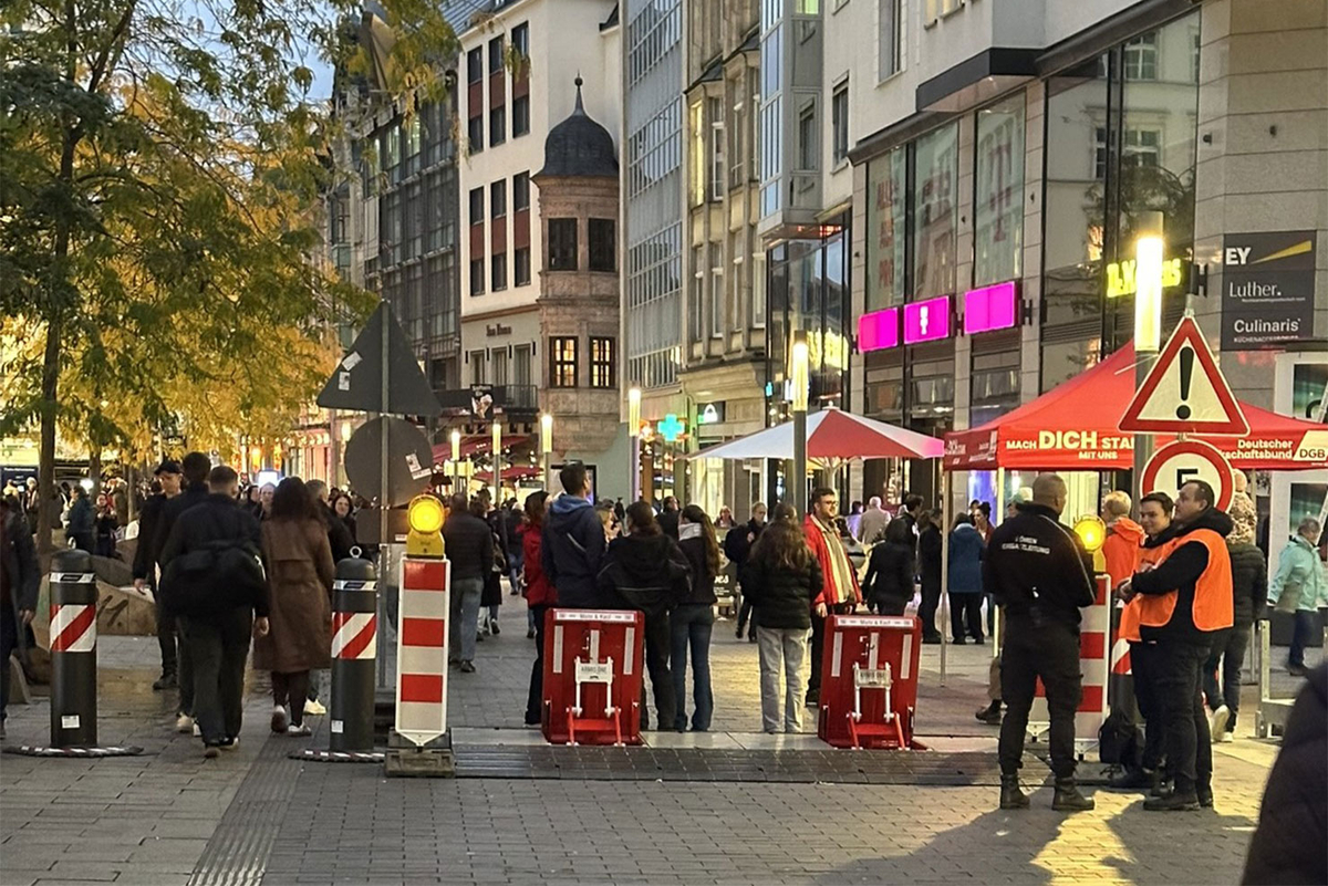 Festbesucher stehen an einer mobilen Fahrzeugsperre in einer Innenstadt